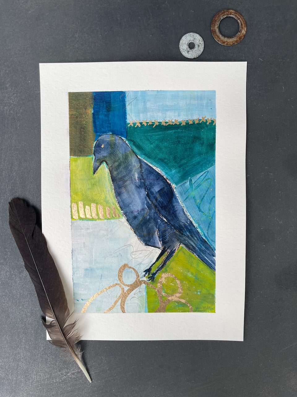 a flat lay photo of a crow painting with a crow feather and two shiny objects