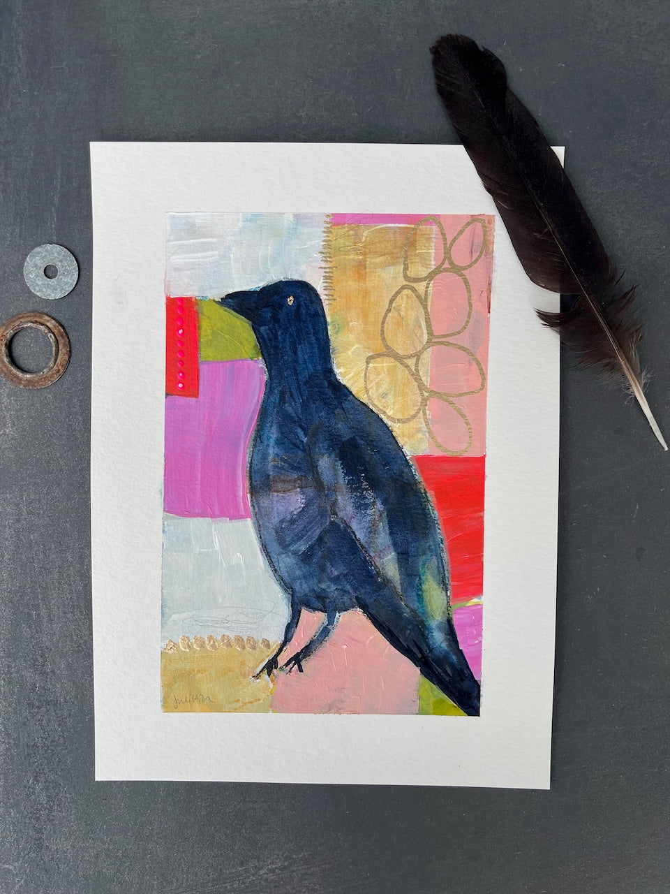 Flat lay of a crow painting embellished with a crow feather and two shiny objects