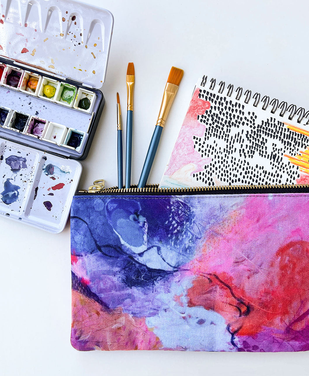 flat lay of brightly colored zipper pouch holding paint brushes and paper with a watercolor paint palette off to the side